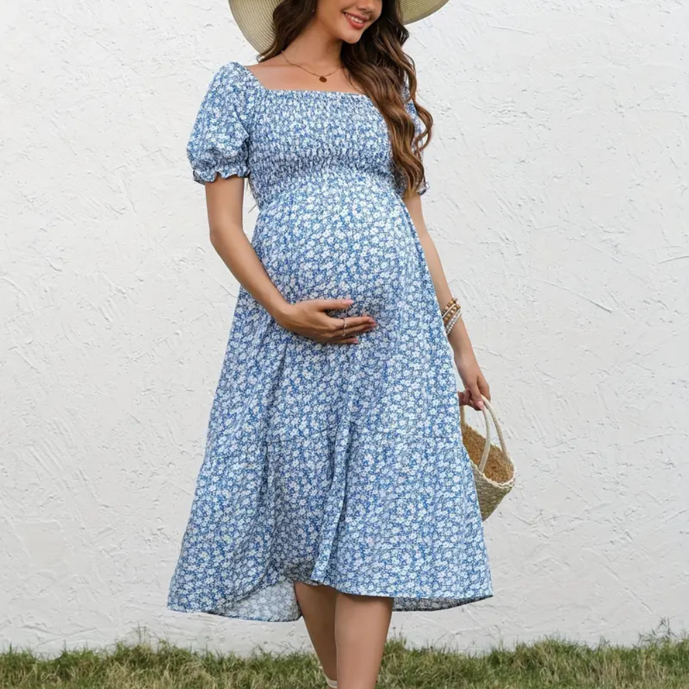 Pregnant woman wearing a blue floral dress against a white wall.