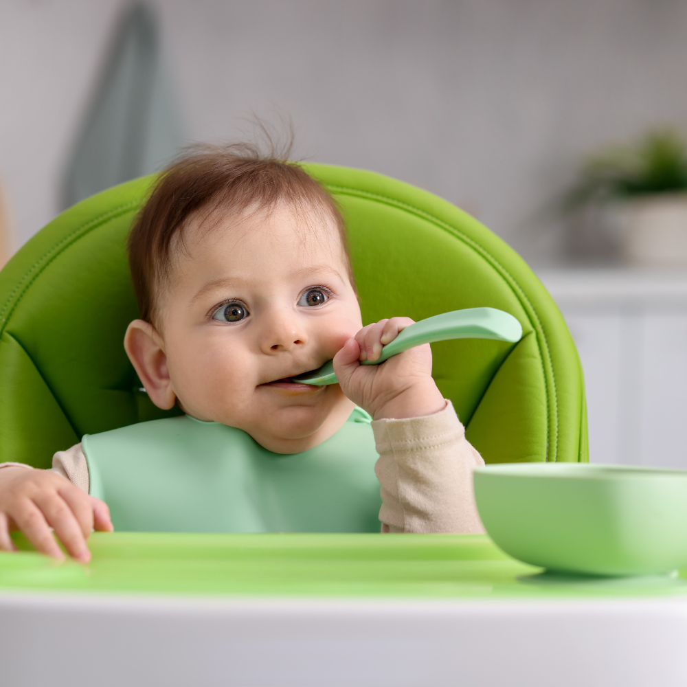 Baby in a green high chair eating with a spoon

