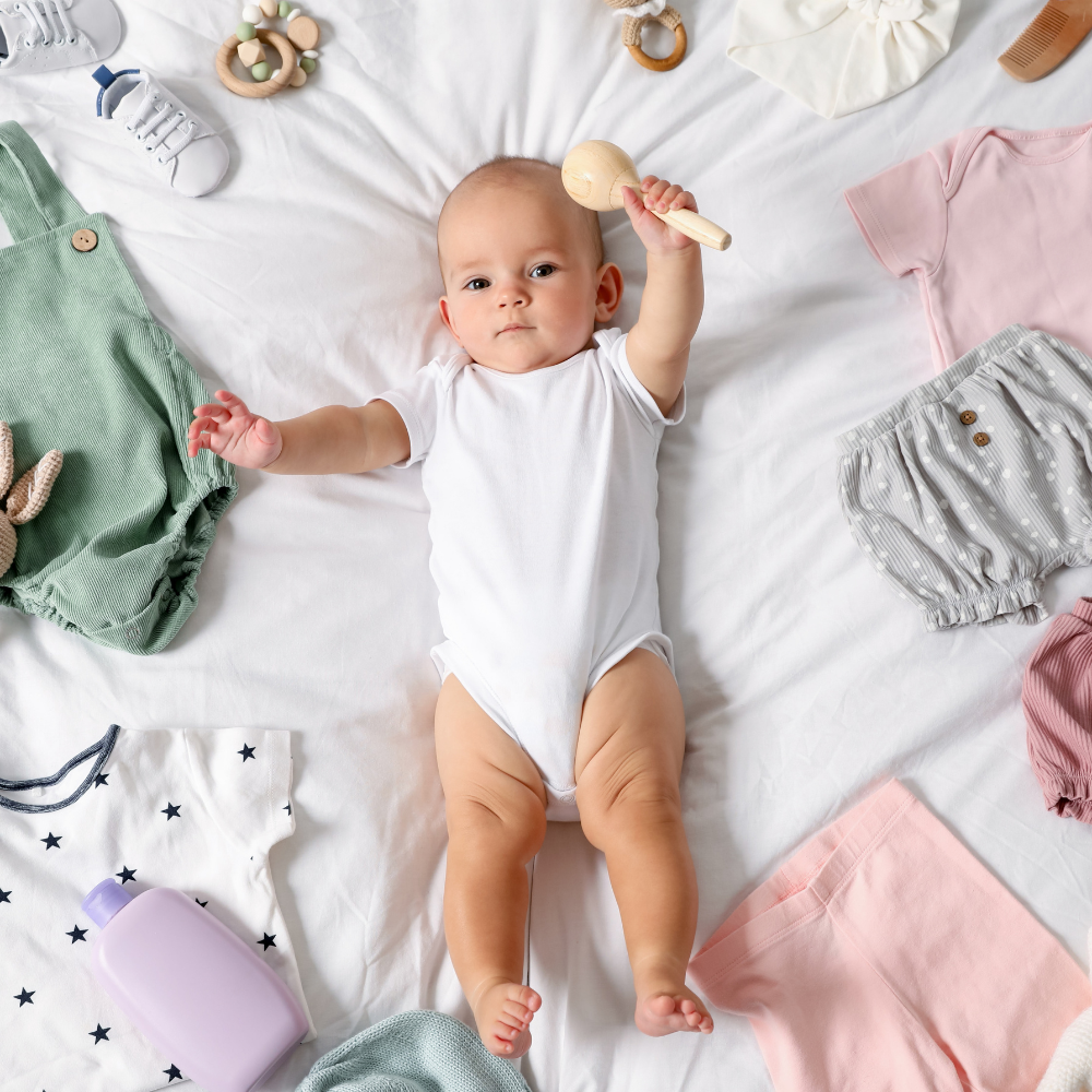 Baby lying on a white blanket surrounded by baby clothes and toys

