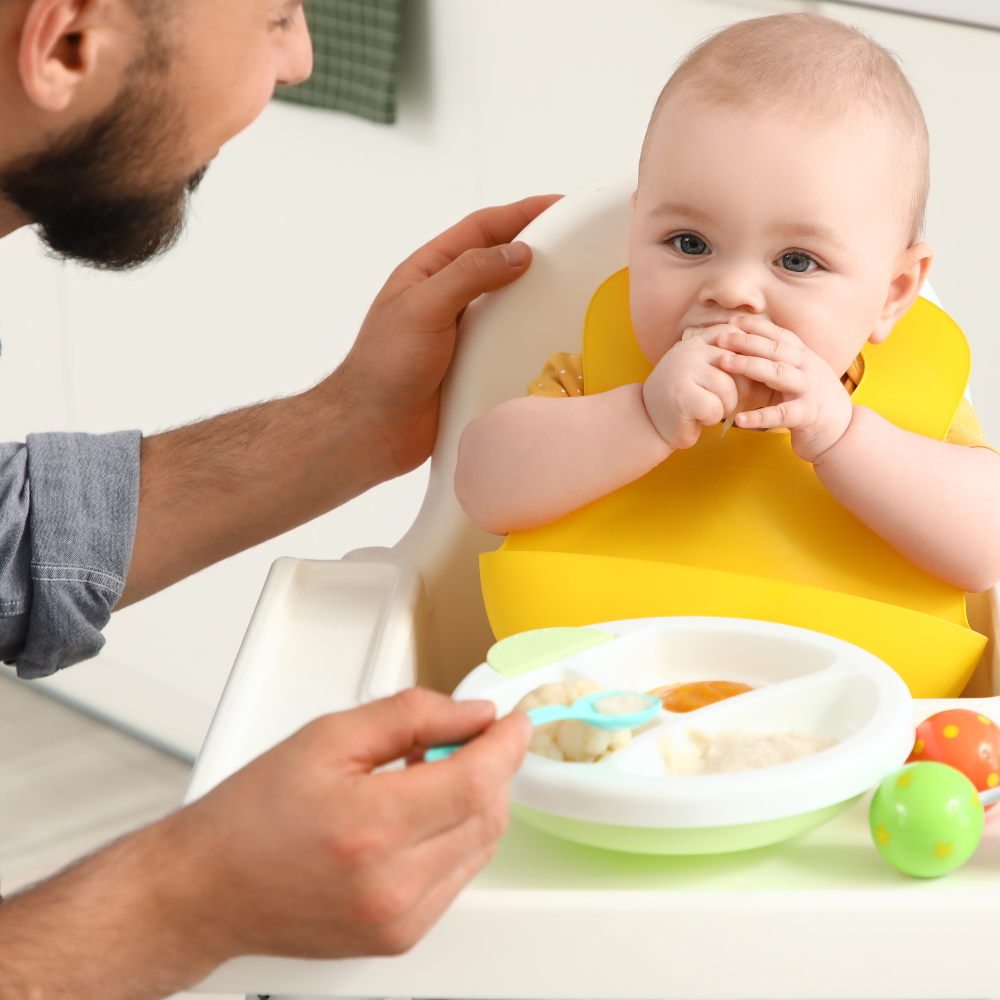 Baby in a yellow bib being fed by an adult in a high chair.

