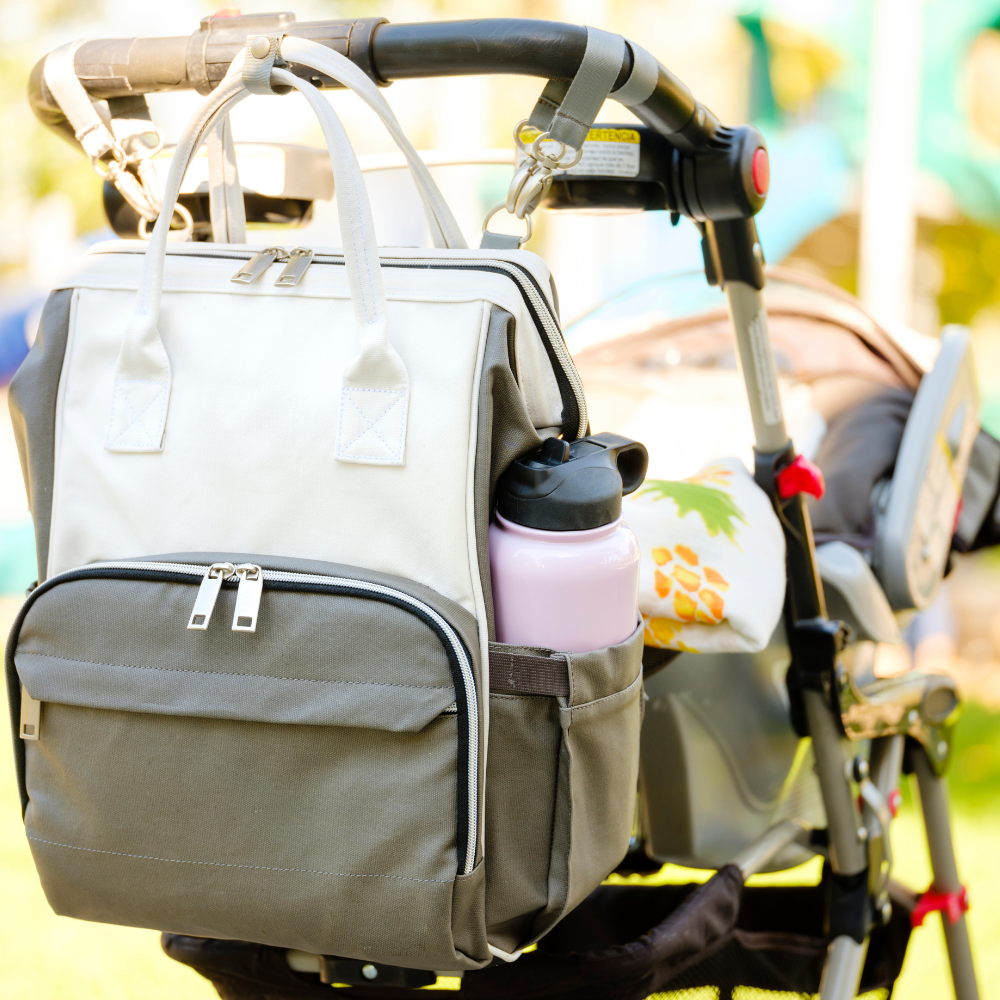 Gray backpack attached to a stroller with a water bottle inside, outdoors.

